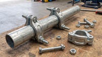 Galvanised drop-forged scaffold right-angle couplers clamped to a 48.3 mm steel scaffold tube on a workbench, with a swivel coupler and loose T-bolts and nuts alongside