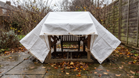 White woven tarpaulin tied with rope over a wooden garden table and chairs on a wet autumn patio, with an air gap left at the bottom